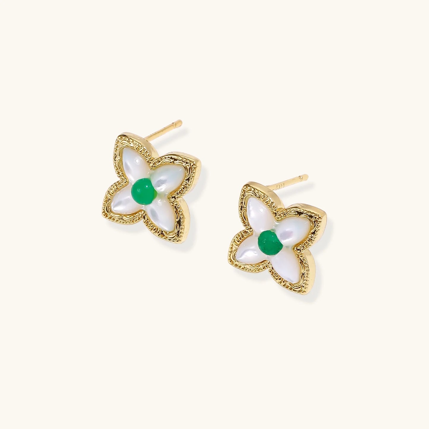 Gold quatrefoil stud earrings with white mother-of-pearl petals and emerald green gemstone center on beige background.