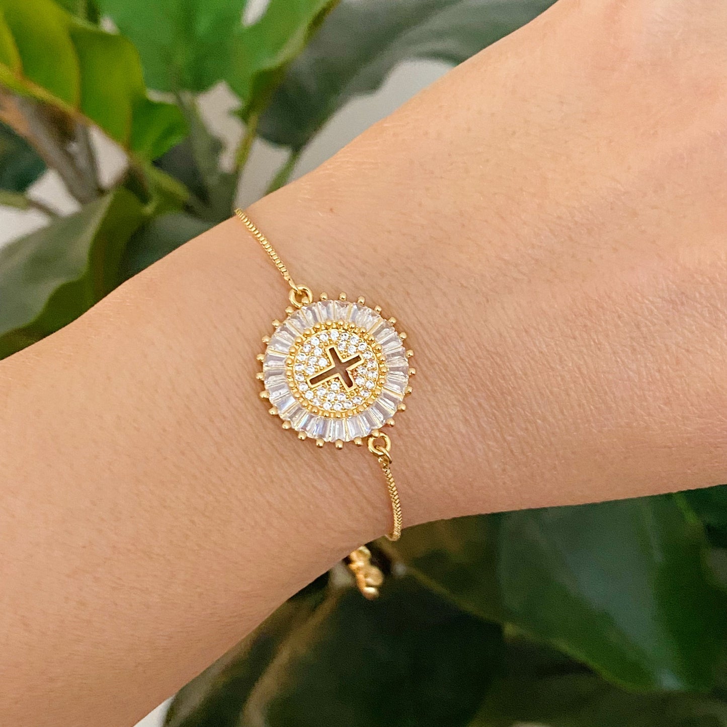 Gold bracelet with a circular design featuring a cross on a wrist against a green leafy background