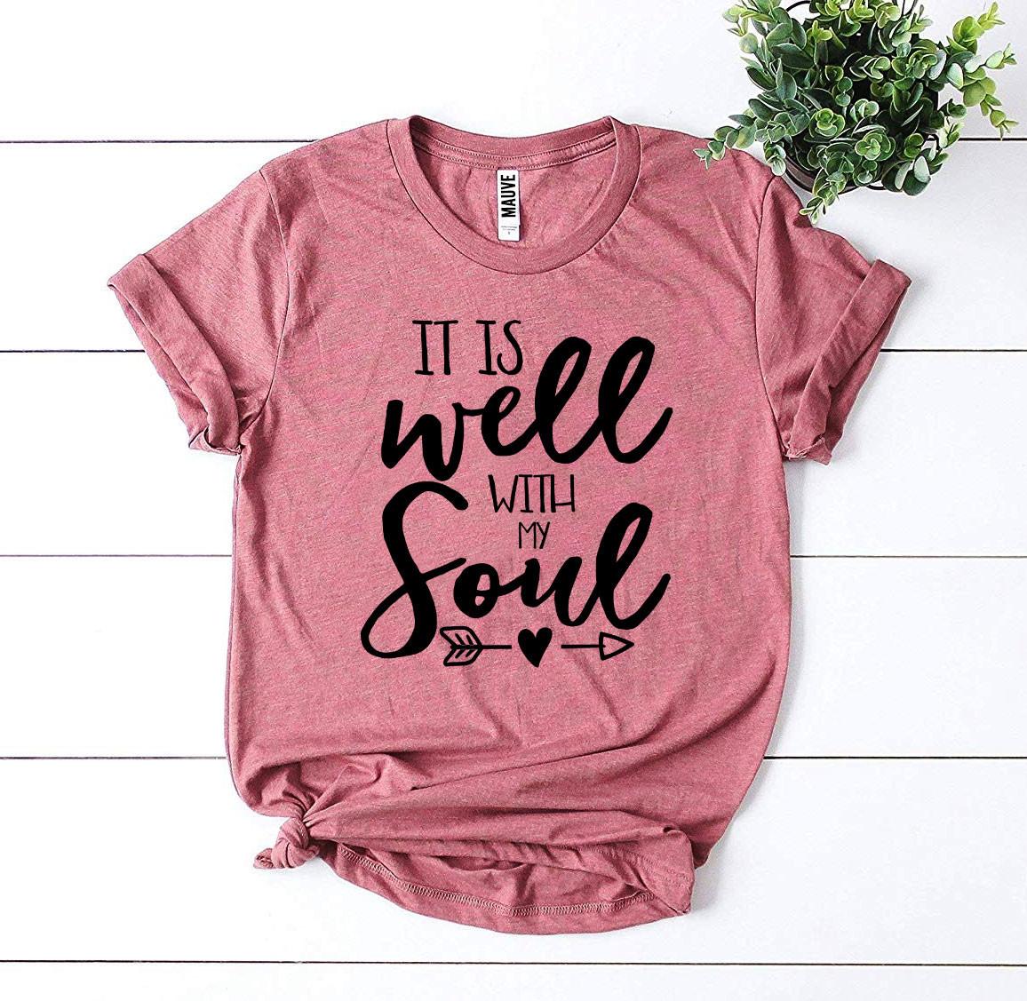 Pink t-shirt with 'It is well with my soul' text on a white wooden surface with a plant.