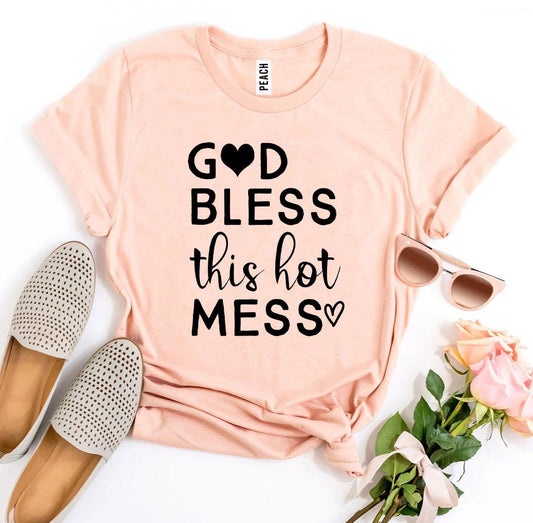 Peach t-shirt with 'God bless this hot mess' text, accompanied by flats, sunglasses, and flowers on a white background.