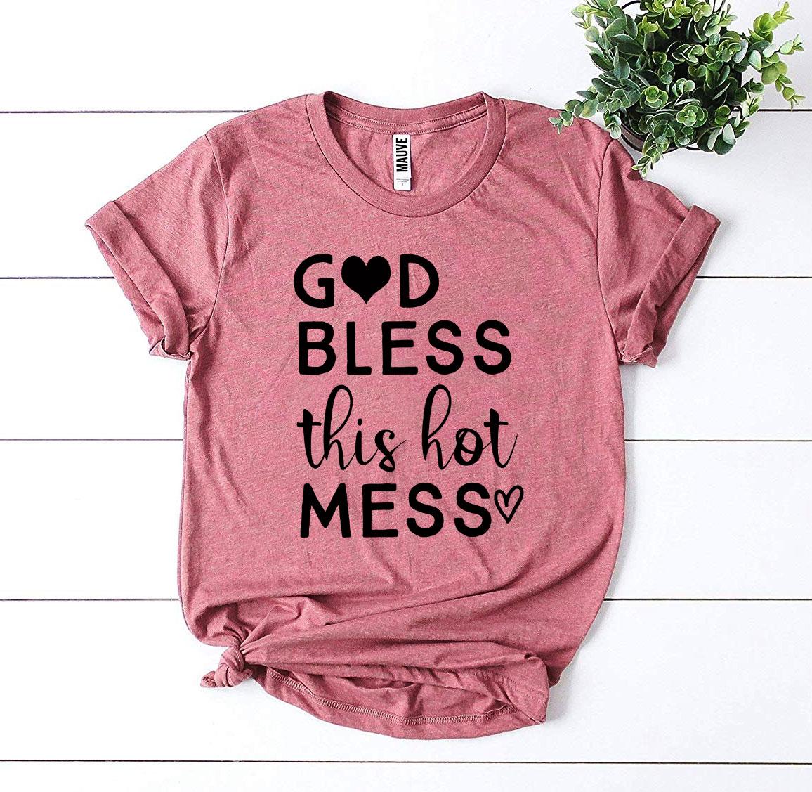 Pink t-shirt with 'God bless this hot mess' text on a white wooden surface with a plant.